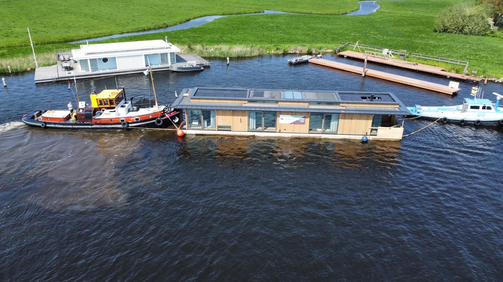 Luchtfoto van een drijvende woning die met een sleepboot aankomt bij een ligplaats in de polder