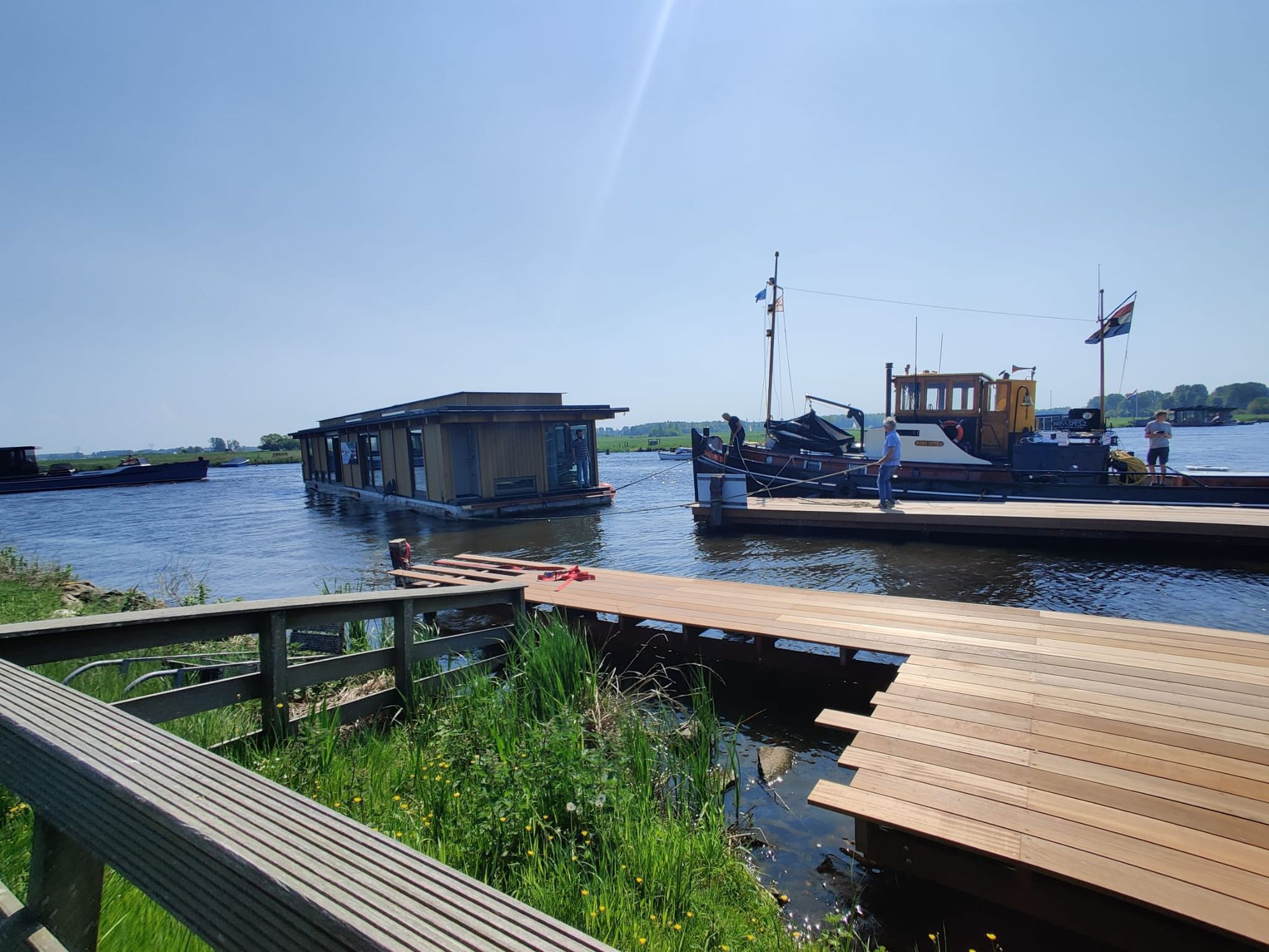 Nieuwe houten steiger aan het water, met een drijvend gebouw en een sleepboot op de achtergrond