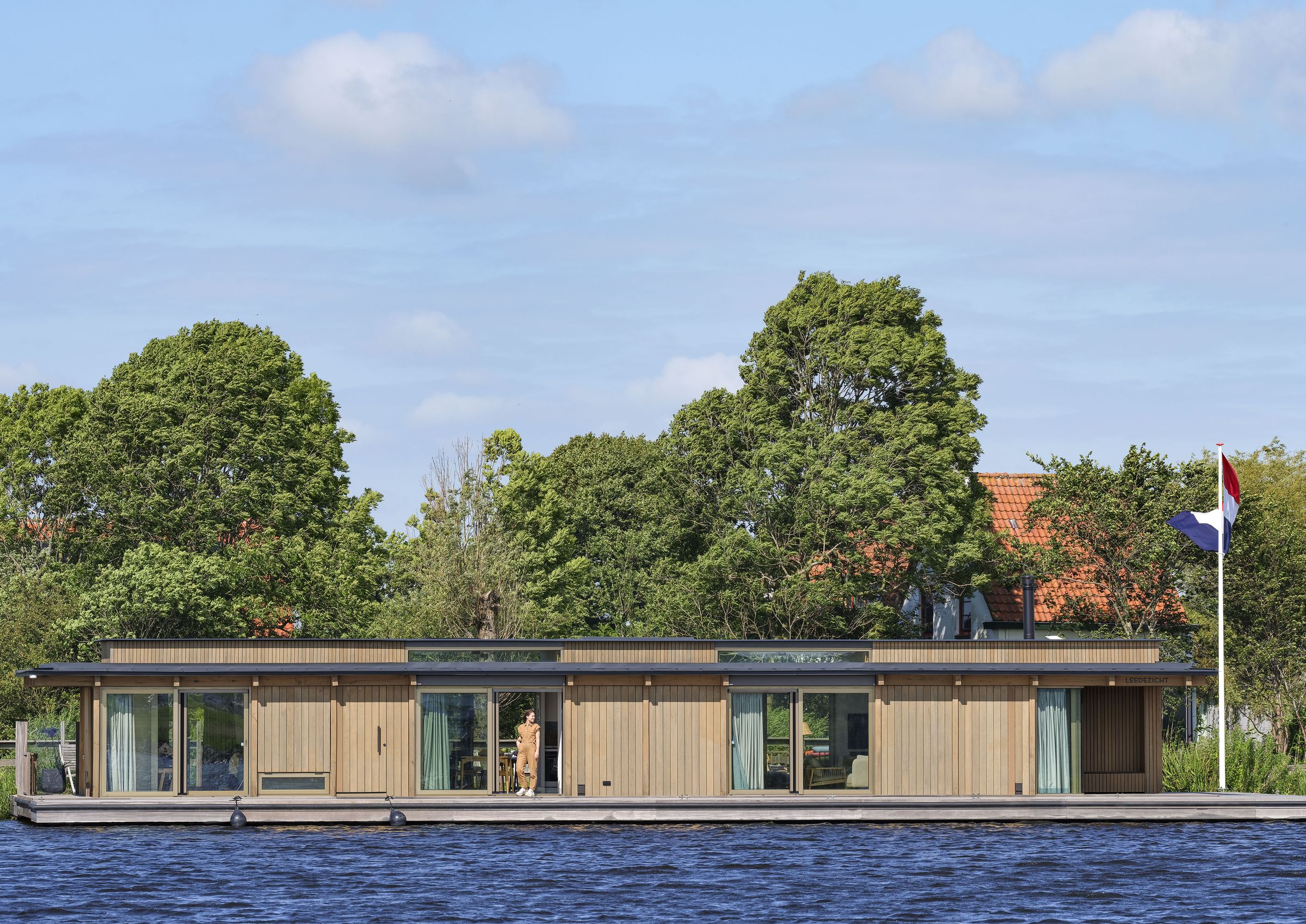 Houten voorgevel van een drijvende woning in Warmond, met bomen en een boerderij erachter