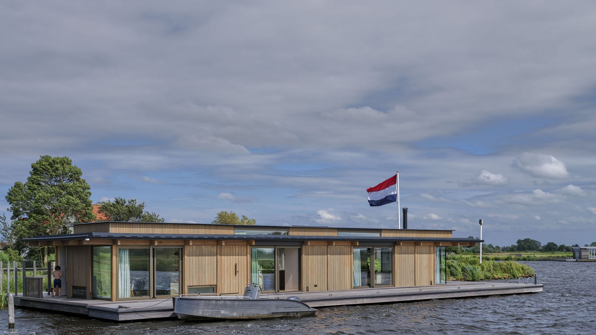 Drijvende woning van Domera met houten gevel en een vlaggenmast, met een sloep langszij